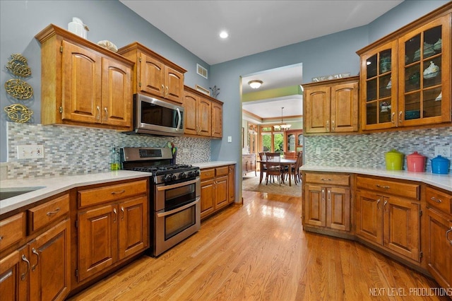 kitchen featuring stainless steel appliances, brown cabinetry, light countertops, light wood-style floors, and glass insert cabinets