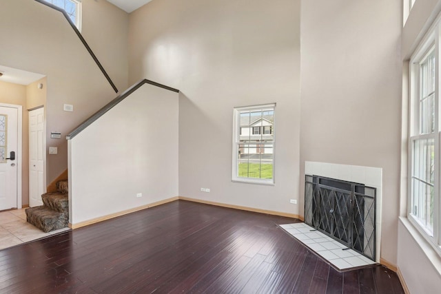 unfurnished living room with a towering ceiling, stairway, wood-type flooring, a tile fireplace, and baseboards