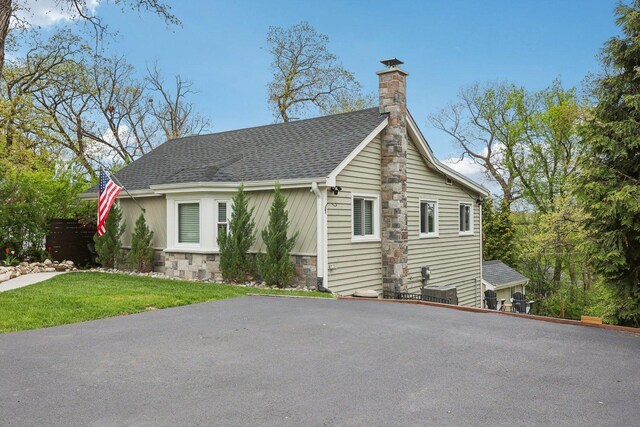 view of front facade featuring stone siding, a shingled roof, a front lawn, and a chimney