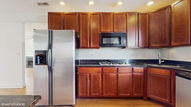 kitchen with appliances with stainless steel finishes, a sink, recessed lighting, light wood-type flooring, and dark stone countertops