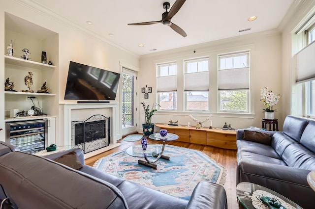 living area with wine cooler, light wood-type flooring, a ceiling fan, ornamental molding, and a fireplace with flush hearth