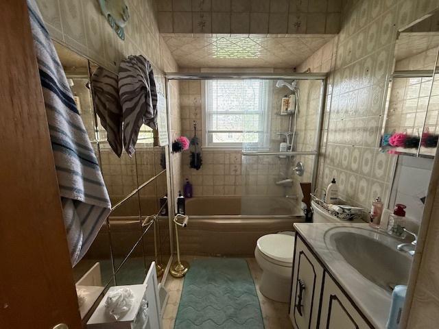 bathroom featuring combined bath / shower with glass door, toilet, tile patterned floors, vanity, and tile walls