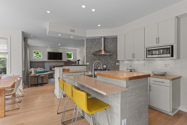 kitchen with stainless steel microwave, wall chimney range hood, light wood-style flooring, backsplash, and plenty of natural light