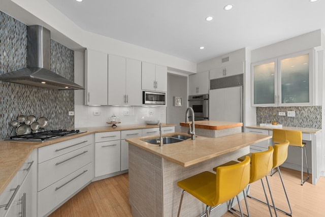 kitchen featuring wall chimney exhaust hood, built in appliances, tasteful backsplash, a sink, and a kitchen bar
