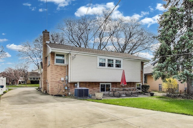 back of property with a chimney, brick siding, central AC unit, a lawn, and a patio