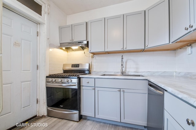kitchen with stainless steel appliances, a sink, under cabinet range hood, light wood-style flooring, and decorative backsplash