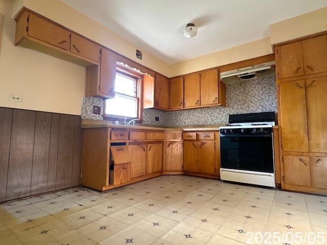 kitchen with under cabinet range hood, brown cabinetry, light floors, range with gas stovetop, and decorative backsplash