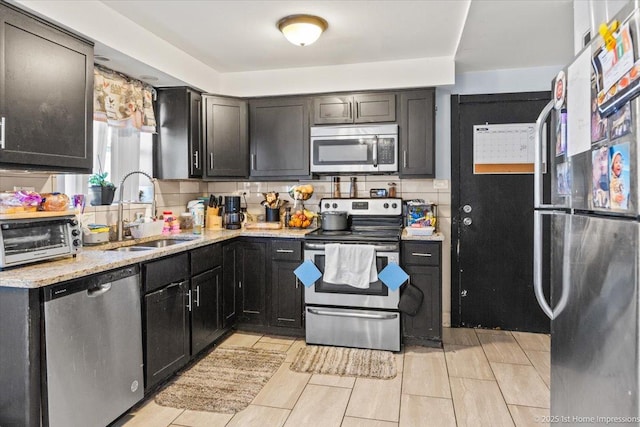 kitchen featuring stainless steel appliances, a sink, light stone counters, tasteful backsplash, and dark cabinets