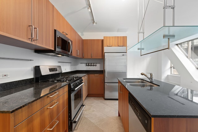 kitchen featuring stainless steel appliances, a sink, brown cabinetry, dark stone countertops, and a center island with sink