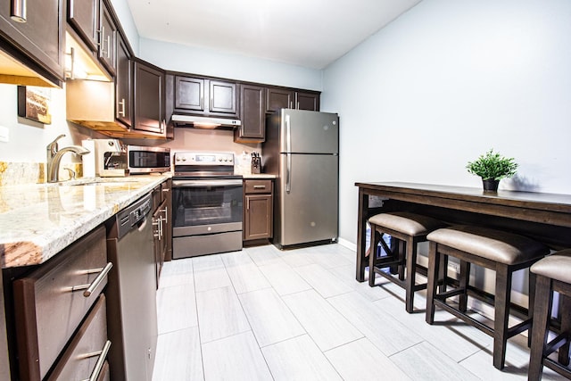 kitchen featuring appliances with stainless steel finishes, dark brown cabinetry, under cabinet range hood, light stone countertops, and a sink