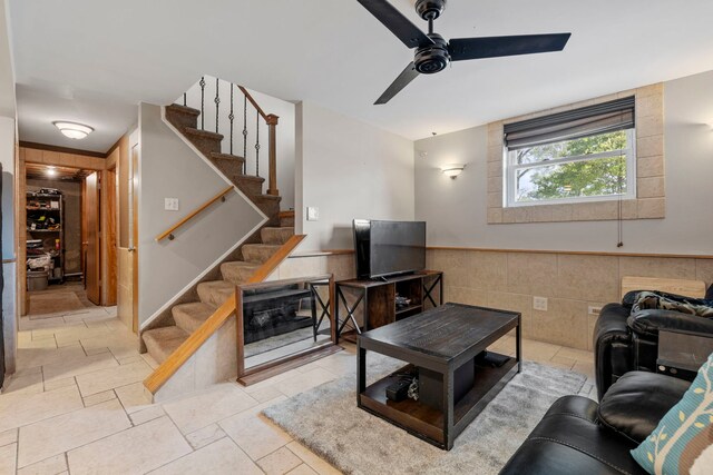 living area with stairs, a ceiling fan, wainscoting, and tile walls