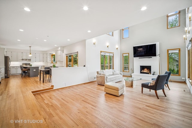 living room with a brick fireplace, light wood finished floors, recessed lighting, and a towering ceiling