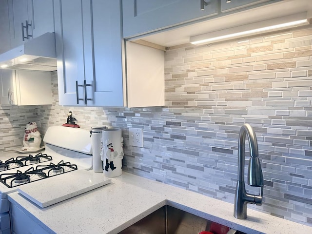 kitchen with white range with gas stovetop, under cabinet range hood, backsplash, light stone countertops, and blue cabinetry