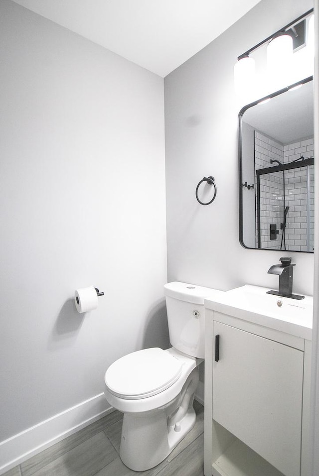 bathroom with toilet, baseboards, vanity, tiled shower, and wood finished floors