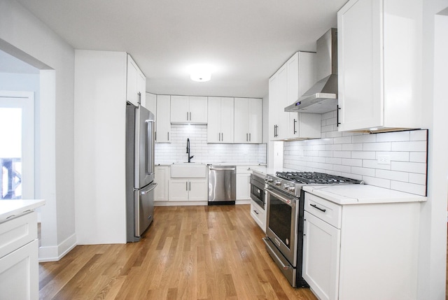 kitchen featuring stainless steel appliances, wall chimney range hood, decorative backsplash, light countertops, and light wood finished floors