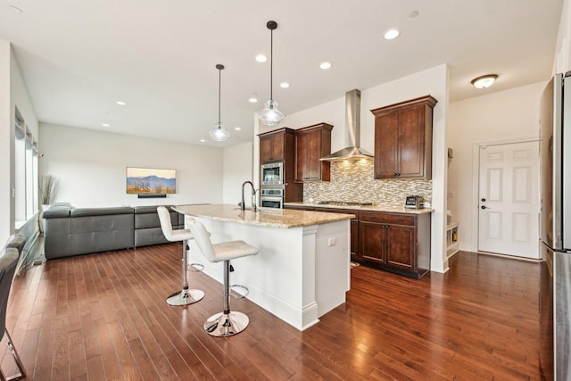 kitchen featuring wall chimney range hood, appliances with stainless steel finishes, open floor plan, dark wood finished floors, and a kitchen breakfast bar