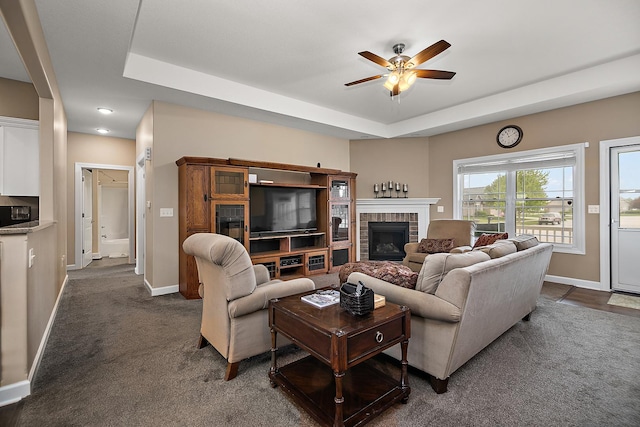 living room featuring a ceiling fan, a raised ceiling, baseboards, a brick fireplace, and dark colored carpet