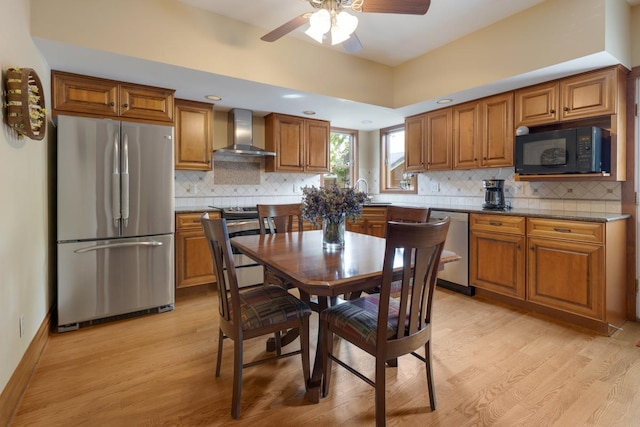 kitchen featuring appliances with stainless steel finishes, wall chimney exhaust hood, brown cabinetry, light wood finished floors, and a ceiling fan