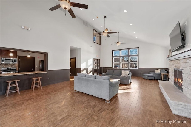 living area with a ceiling fan, a stone fireplace, wainscoting, high vaulted ceiling, and wood finished floors