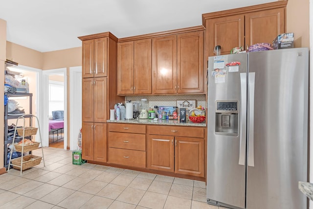 kitchen with stainless steel refrigerator with ice dispenser, brown cabinetry, decorative backsplash, light tile patterned floors, and light stone countertops