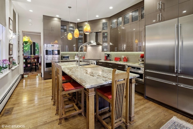 kitchen with appliances with stainless steel finishes, a baseboard heating unit, wall chimney range hood, a sink, and light wood-style floors