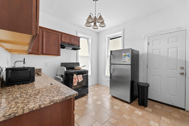 kitchen with freestanding refrigerator, black gas range, under cabinet range hood, a chandelier, and light stone finish flooring