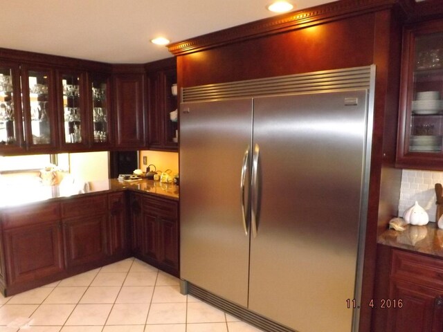 kitchen with built in refrigerator, glass insert cabinets, light tile patterned floors, tasteful backsplash, and reddish brown cabinets