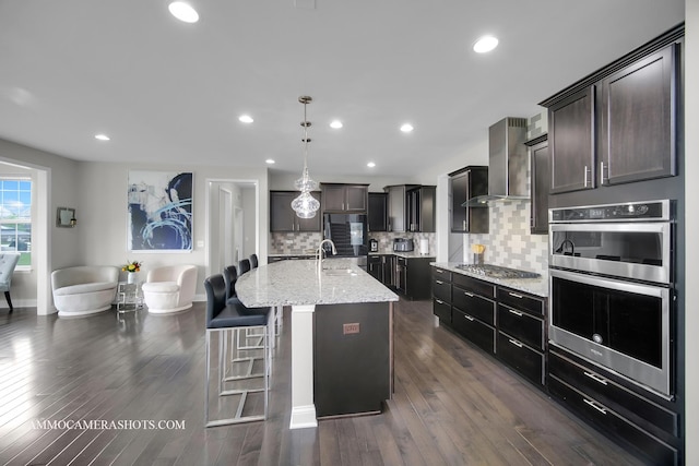 kitchen featuring wall chimney exhaust hood, tasteful backsplash, double oven, light stone countertops, and a kitchen island with sink