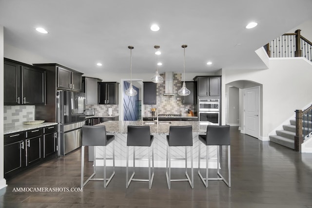 kitchen with stainless steel appliances, wall chimney range hood, arched walkways, light stone countertops, and backsplash