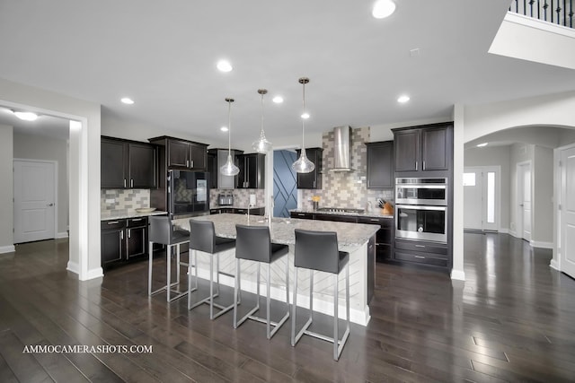 kitchen with wall chimney exhaust hood, arched walkways, stainless steel appliances, tasteful backsplash, and a center island with sink