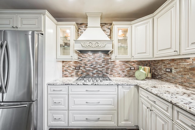 kitchen with stainless steel appliances, custom range hood, backsplash, light stone countertops, and glass insert cabinets