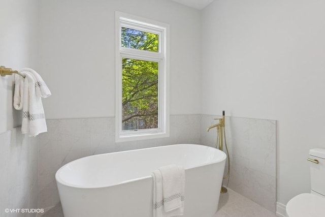 full bath featuring tile walls, toilet, a freestanding tub, a wainscoted wall, and tile patterned floors