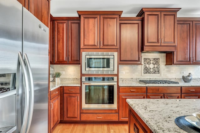 kitchen featuring stainless steel appliances, light stone counters, backsplash, light wood-style flooring, and brown cabinets