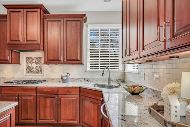 kitchen featuring a sink, stainless steel gas cooktop, tasteful backsplash, light stone counters, and reddish brown cabinets