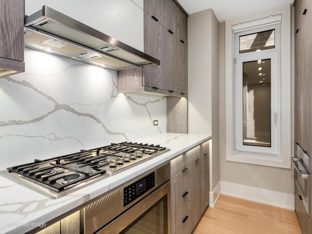 kitchen with wall chimney range hood, stainless steel gas cooktop, light wood-style flooring, baseboards, and light stone counters