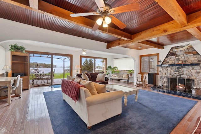 living area featuring beamed ceiling, wooden ceiling, ceiling fan, a stone fireplace, and hardwood / wood-style flooring