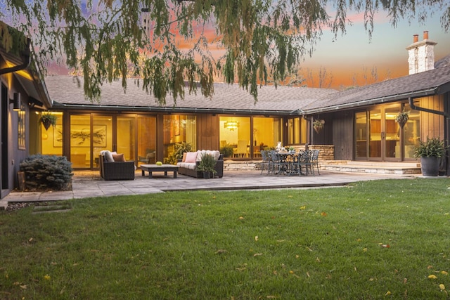 back of house at dusk featuring an outdoor living space, a chimney, a lawn, a patio, and roof with shingles