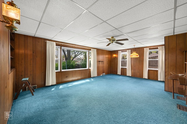 unfurnished living room featuring carpet flooring, healthy amount of natural light, a ceiling fan, wooden walls, and a drop ceiling