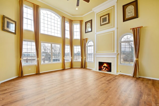 unfurnished living room featuring ceiling fan, baseboards, light wood-style floors, ornamental molding, and a towering ceiling