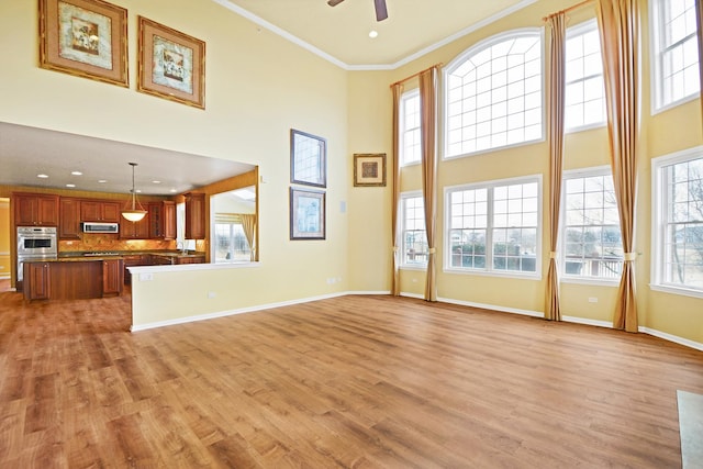 unfurnished living room featuring ceiling fan, light wood-type flooring, a towering ceiling, ornamental molding, and baseboards