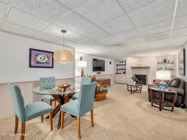 carpeted dining space with built in shelves, a brick fireplace, wainscoting, and a paneled ceiling