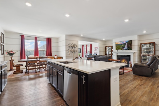 kitchen featuring stainless steel dishwasher, a sink, light countertops, a center island with sink, and dark wood-style flooring