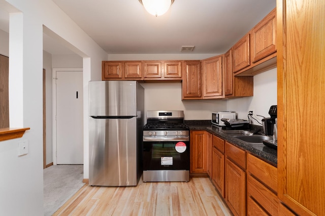 kitchen featuring appliances with stainless steel finishes, a sink, dark countertops, light wood-type flooring, and brown cabinets