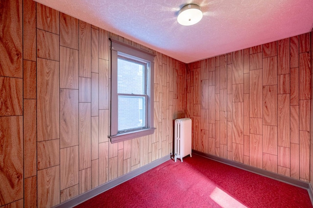 carpeted empty room with radiator, a textured ceiling, wood walls, and baseboards