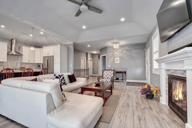 living room with ceiling fan, light wood finished floors, baseboards, a glass covered fireplace, and a tray ceiling