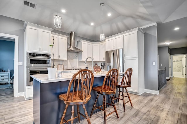 kitchen featuring wall chimney range hood, light countertops, appliances with stainless steel finishes, light wood finished floors, and a center island with sink