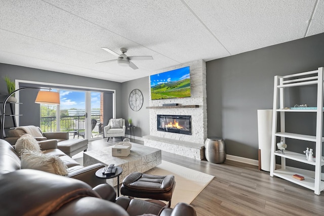 living area featuring ceiling fan, a stone fireplace, wood finished floors, baseboards, and a textured ceiling
