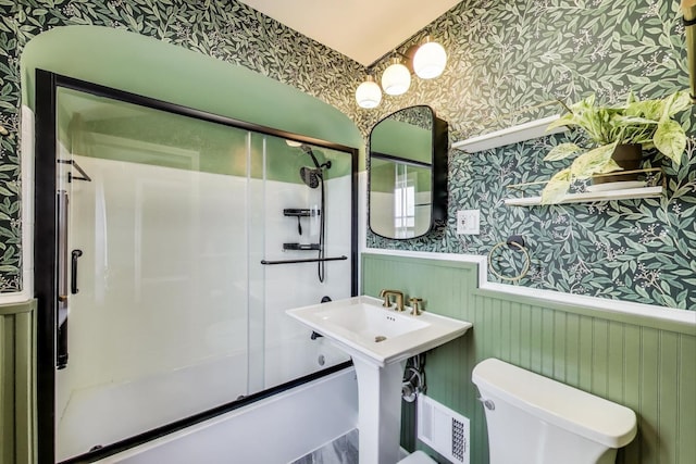 full bathroom with wallpapered walls, a wainscoted wall, toilet, and shower / bath combination with glass door
