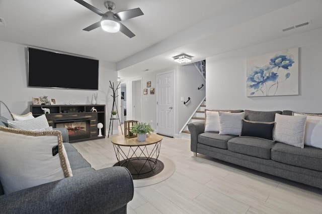 living room featuring stairway, wood finished floors, a ceiling fan, and a glass covered fireplace