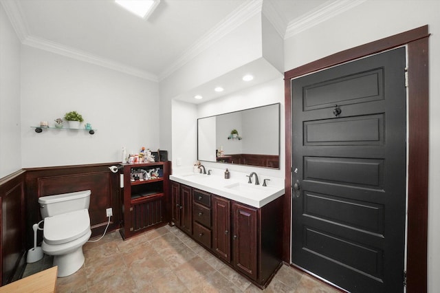 bathroom with toilet, crown molding, double vanity, and a wainscoted wall